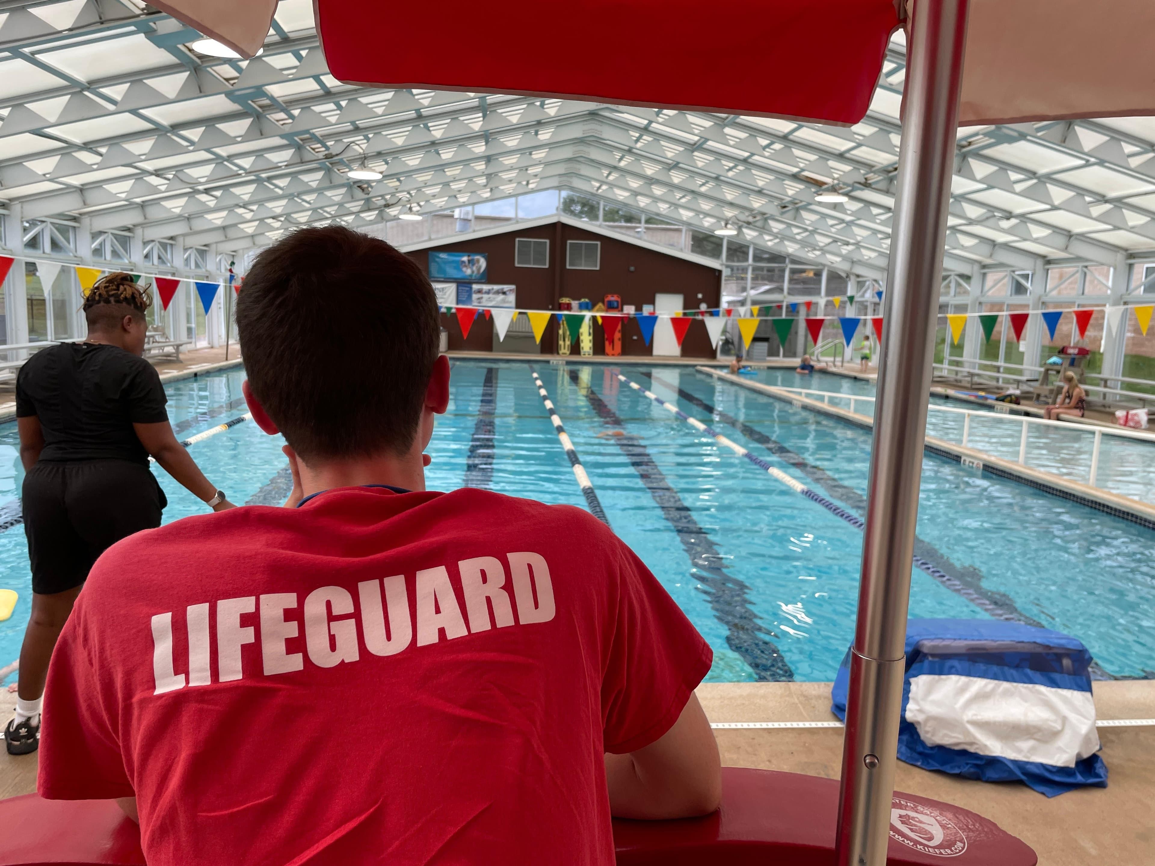 A lifeguard in a red LIFEGUARD shirt watching an indoor pool decorated with pennant flags.