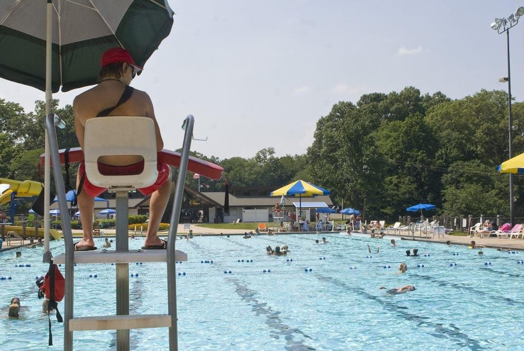A lifeguard in a red cap seen from behind on a guard chair, watching a busy outdoor pool.