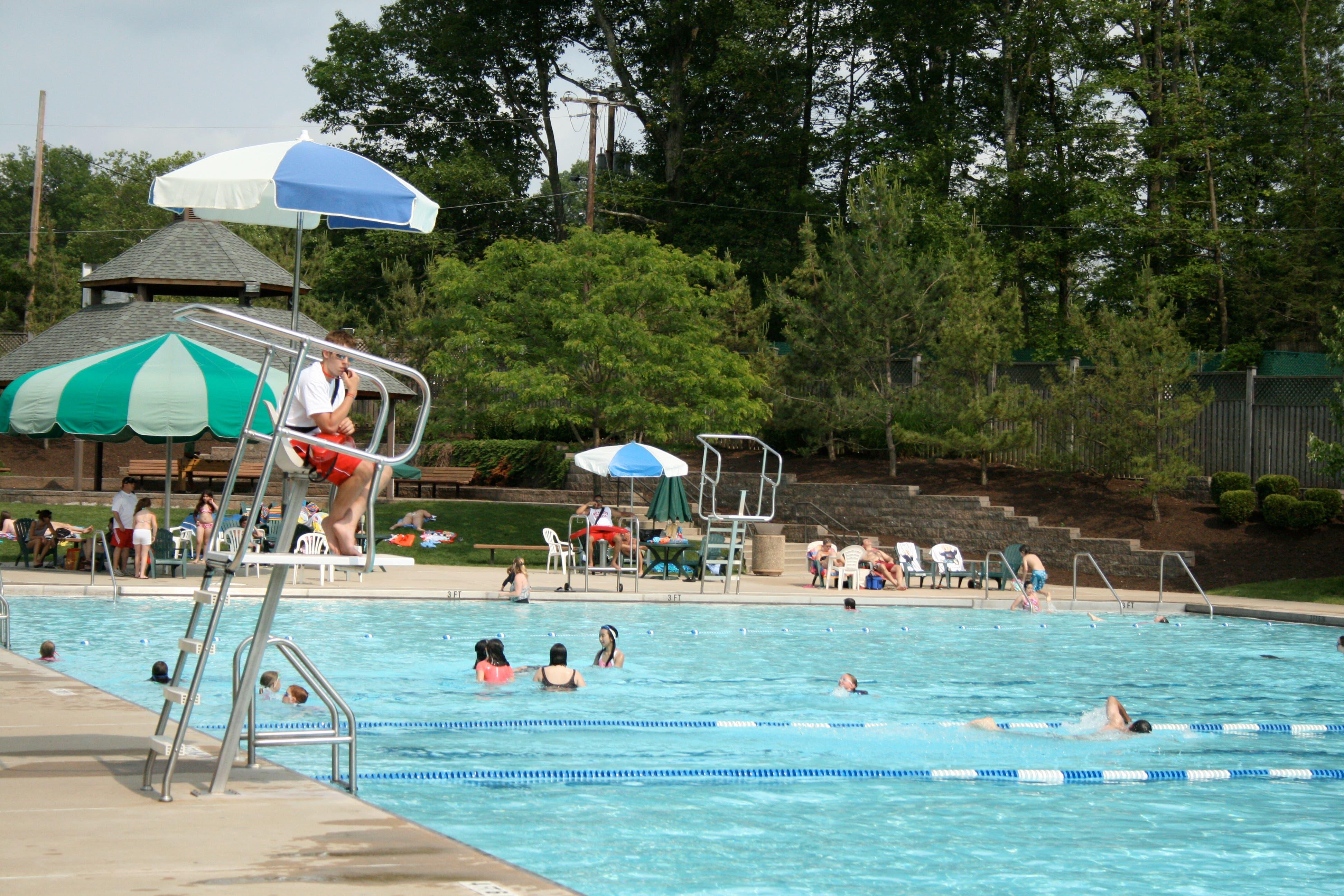 A lifeguard on a tall guard stand under an umbrella, watching swimmers in a large community pool surrounded by trees.