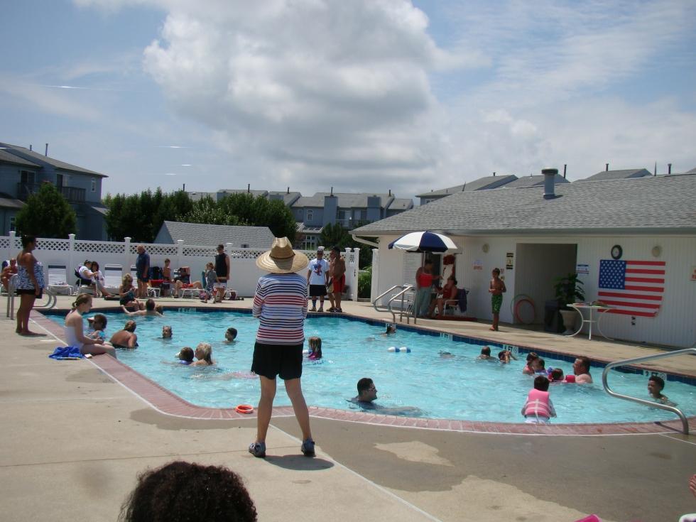 A staff member in a sun hat watching children swim in a community pool beside an American flag.